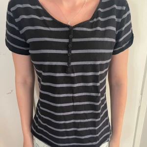 Svart och grå randig t-shirt med knappar - Snygg svart t-shirt med gråa horisontella ränder och korta ärmar. Framtill finns en knappslå med fem svarta knappar och en lätt v-ringad hals. T-shirten har en normal passform och är gjord i mjuk bomull, perfekt för en chill och avslappnad stil.