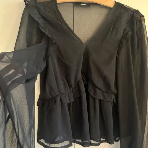 Svart blus med volanger från Bikbok - Säljer en svart blus från Bikbok i storlek M. Blusen har transparenta, långärmade ärmar och snygga volangdetaljer längs axlar och midja. V-ringad hals och lätt, flowy material som ger en cool vibe.  