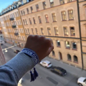 Blå armband från Jacob Cohën - Snyggt blått armband från Jacob Cohën med vit text och detaljer. Armbandet har ett randigt mönster med små vita prickar längs kanterna och avslutas med coola blå tofsar. Perfekt accessoar för att ge din outfit lite extra edge. 