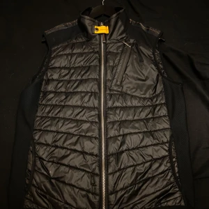 Svart dunväst från Parajumpers L - Snygg svart dunväst från Parajumpers med quiltad design och cool gul detalj vid kragen. Västen har dragkedja framtill, bröstficka med dragkedja och orange insida. Perfekt lager-på-lager-plagg för kyliga dagar.