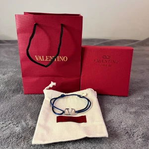 Valentino armband  - Snyggt blått armband från Valentino Garavani med justerbar snörning och silverfärgat V-logga i metall. Armbandet har en tunn, glansig blå tråd och en liten metallbricka med Valentino-gravyr. Perfekt accessoar för att lyfta din stil.