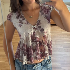 Blommig spetsblus med rosett - Supersöt blommig blus i ljus spets med lila och vinröda toner. Croppad och flowy passform, v-ringad med liten rosett framtill. Ärmlös design och transparent material som ger en romantisk vibe. Perfekt att styla med jeans eller kjol för en trendig look.