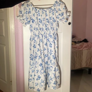 Vit blommig sommarklänning med puffärm - Supersöt vit klänning med blått blommigt mönster och puffiga korta ärmar. Klänningen har smock i midjan och en luftig kjol med volang. Perfekt för varma dagar och har en romantisk vibe med sin fyrkantiga ringning och mjuka bomullskänsla.