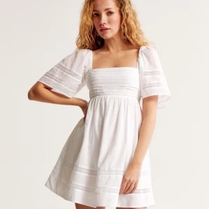 Vit klänning med fyrkantig urringning - Söt vit klänning med fyrkantig urringning och korta ärmar. Den har en luftig passform och fina spetsdetaljer på ärmar och kjol. Perfekt för sommarens varma dagar.