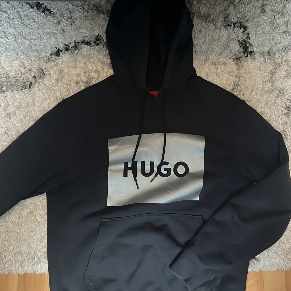 Snygg svart hoodie från Hugo med stor logga på bröstet. Tröjan har en klassisk känguruficka och justerbar huva med dragsko. Perfekt för en avslappnad stil.. Neuletakit & Villapaidat.