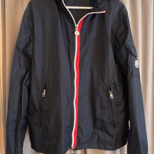 Svart vindjacka från Moncler - Säljer en stilren svart vindjacka från Moncler med röd och vit dragkedja. Jackan har en huva och praktiska fickor med dragkedja. Perfekt för blåsiga dagar och har en snygg logga på ärmen.