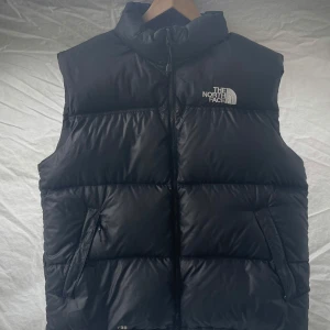 Svart dunväst från The North Face - Säljer en svart dunväst från The North Face med dragkedja och hög krage. Västen har två fickor med dragkedja och logotypen synlig på bröstet och ryggen. Perfekt för kyliga dagar. Västen är knappt använd, ny pris ca 2000kr
