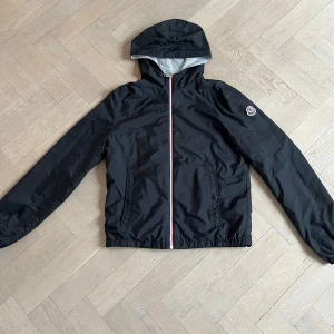 Svart vindjacka från Moncler - Säljer en stilren svart vindjacka från Moncler med huva och dragkedja i storlek 14/164. Jackan har långa ärmar och elastiska muddar vid ärmslut och nederkant. Jackan är köpt på NK i Stockholm i oktober 2024.