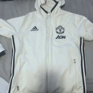 United Vit vindjacka från Adidas - Säljer en vit vindjacka från Adidas med Manchester United-logga och AON-tryck. Jackan har dragkedja och huva, samt svarta detaljer längs ärmarna och sidorna. Perfekt för fotbollsfans!