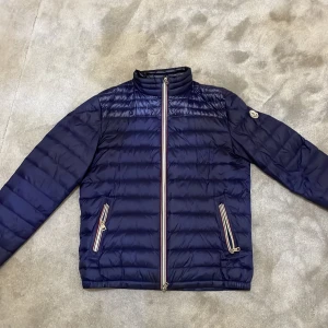 Blå dunjacka från Moncler - Säljer en snygg blå dunjacka från Moncler med dragkedja och två fickor framtill. Jackan har ett stilrent märke på ärmen och är perfekt för kyligare dagar. Den är långärmad och har en klassisk passform. Priset kan diskuteras!!!