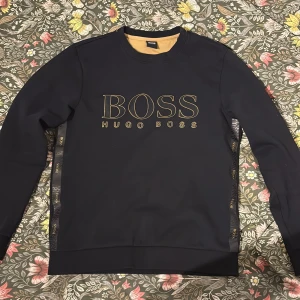 Svart tröja från Hugo Boss - Snygg svart tröja från Hugo Boss med deras logga i guld på bröstet. Tröjan har långa ärmar och ribbade muddar vid ärmslut och nederkant. Perfekt för en stilren look.
