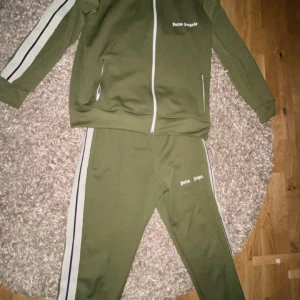 Olivgrön träningsset från Palm Angels - Säljer ett olivgrönt dress från Palm Angels med vita och svarta ränder längs ärmar och ben. Setet har dragkedja framtill och är perfekt för en sportig look. Det är tillverkat i ett mjukt och bekvämt material ny tvättad och är äkta finns ej kvitto. Pris kan även sänkas vid mer information kontakta mig.