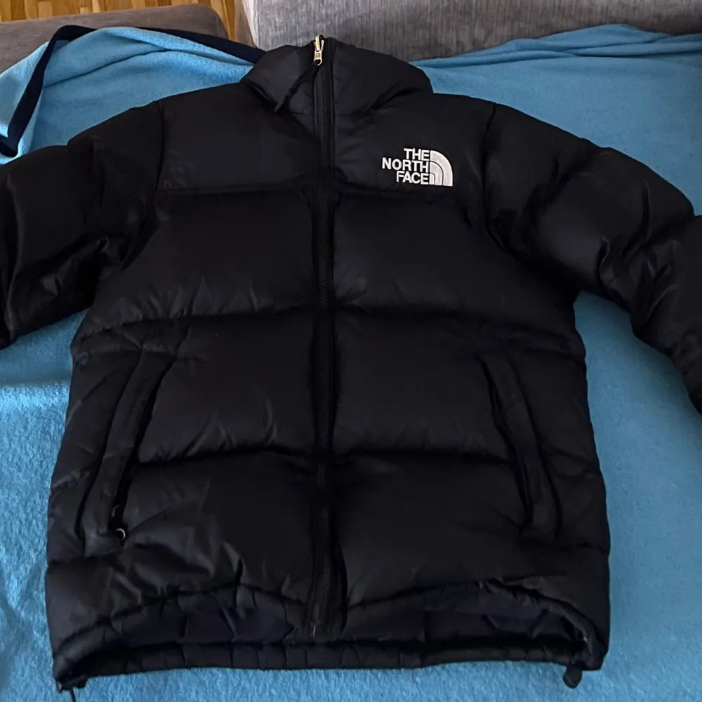 Säljer en svart dunjacka från The North Face med dragkedja och logotyp på bröstet. Jackan har en pufferdesign och är perfekt för kalla dagar. Den är långärmad och har en hög krage för extra värme.. Takit.
