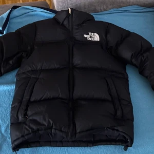 Svart dunjacka från The North Face - Säljer en svart dunjacka från The North Face med dragkedja och logotyp på bröstet. Jackan har en pufferdesign och är perfekt för kalla dagar. Den är långärmad och har en hög krage för extra värme.