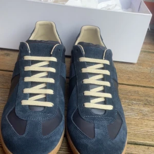 Blå sneakers från Maison Margiela - Snygga blå sneakers från Maison Margiela med en stilren design. Skorna har en ovandel i mocka och skinn, med vita snören och en gummisula. Perfekta för en trendig look. Storlek 41 men sitter som 42 och såklart äkta all denns tillbehör följer med alltså box och dustbag. Ny tvättade. Pris ej hugget i en sten