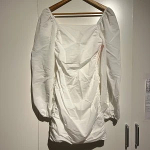 Vit klänning med puffärmar - Snygg vit klänning med puffiga långärmar och rynkdetaljer. Perfekt för en stilren look. Klänningen har en dragkedja i ryggen för enkel påklädning. Aldrig använd. Jätte skönt material 