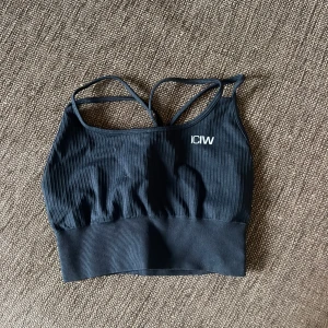 Svart ribbad sporttopp från ICANIWILL - Säljer en svart ribbad sporttopp från ICANIWILL med smala axelband och brottarrygg. Perfekt för träning och ger bra stöd. Toppen är i ett mjukt och stretchigt material för maximal komfort.