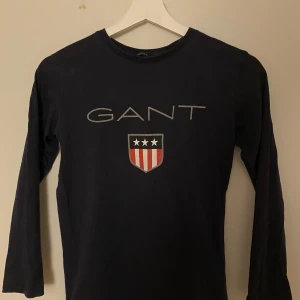 Mörkblå långärmad tröja från GANT - Säljer en svart långärmad tröja från GANT med ett tryck av märkesnamnet och en sköld med stjärnor och ränder på bröstet. Perfekt för en stilren och avslappnad look.