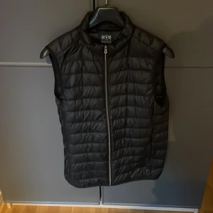 Svart dunväst från BOGI - Snygg svart dunväst från BOGI med dragkedja framtill. Perfekt för lager-på-lager under kyliga dagar. Västen har en stilren design och är lätt att matcha med olika outfits. 