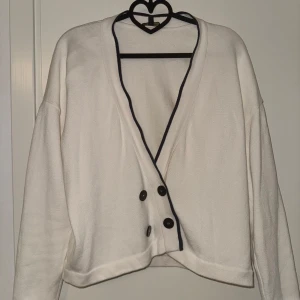 Vit kofta med knappar ifrån Massimo dutti - Säljer en stilren vit kofta med svarta knappar och kontrasterande mörkblå kanter. Koftan har en v-ringad design och långa ärmar, perfekt för en avslappnad look. Passar bra till både jeans och kjol. Perfekt för kyliga dagar!