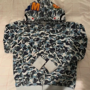 Camouflage hoodie från A Bathing Ape - Säljer en cool camouflage hoodie från A Bathing Ape med dragkedja och detaljer i blått, grönt och svart. Hoodien har en unik design på huvan. Storlek L, öppen för prisförslag, har fler bilder så kontakta om frågor! AA version 
