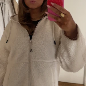 Vit teddyjacka med dragkedja - Mysig vit teddyjacka med dragkedja och huva. Perfekt för kyliga dagar. Jackan har en avslappnad passform och långa ärmar.