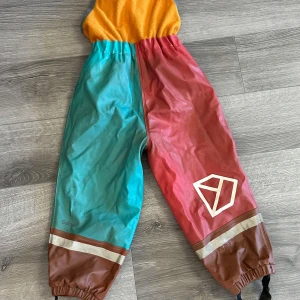 Färgglad regnoverall från Didriksons - Säljer en färgglad regnoverall från Didriksons i Galon-material. Overallen har en mix av turkos, röd och gul färg med bruna detaljer. Den är 100% vattentät och vindtät, perfekt för regniga dagar. Overallen har en dragkedja framtill och förstärkta knän för extra hållbarhet.