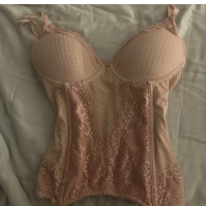 Rosa spetskorsett - Elegant rosa korsett med spetsdetaljer och prickigt mönster. Den har justerbara axelband och hakknäppning bak. Perfekt för en stilfull look.