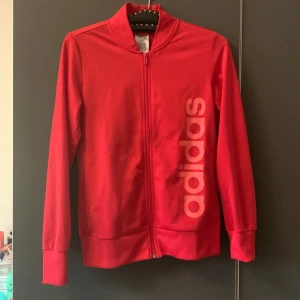Rosa-röd träningsjacka från Adidas - Säljer en snygg rosa-röd träningsjacka från Adidas med dragkedja framtill och stort tryck på sidan. Jackan har långa ärmar och en bekväm passform, perfekt för sport och fritid. Passar både för träning och som en stilren vardagsjacka.🩷