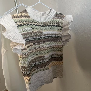 Mönstrad stickad topp från Zara - Säljer en unik stickad topp från Zara med ett färgglatt mönster i grönt, brunt och beige. Toppen har volangdetaljer vid ärmarna och en ribbad rund halsringning. Perfekt för att ge din outfit en bohemisk touch.
