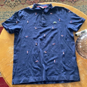 Blå pikétröja från Lacoste - Säljer en snygg blå pikétröja från Lacoste med broderade detaljer i rött och vitt. Tröjan har korta ärmar och en klassisk krage med knappar. Perfekt för en stilren look!