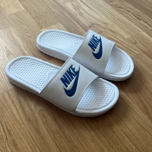 Vita sandaler från Nike - Säljer ett par vita Nike sandaler med blå logga. Perfekta för sommardagar och avslappnade tillfällen. Sandalerna har en bekväm sula och en bred rem över foten med Nike-logotypen i blått.