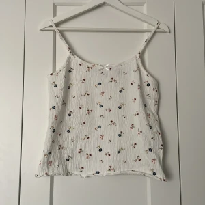 Vit blommig linnetopp - Söt vit linnetopp med små blommor i rosa och blått. Toppen har smala axelband och en liten rosett framtill. Perfekt för varma dagar!