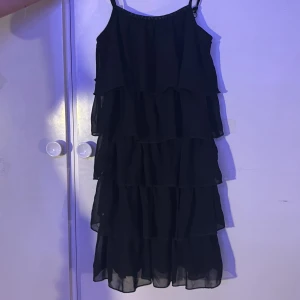 Svart axelbandslös klänning med volanger - Snygg svart klänning med flera lager av volanger och smala axelband. Klänningen har en dekorativ dragkedja på baksidan och glittrande detaljer vid halsringningen. Perfekt för en kväll ute!