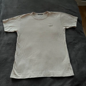 Beige t-shirt från Acne Studios - Säljer en stilren beige t-shirt från Acne Studios. T-shirten har en enkel design med en liten fyrkantig patch på bröstet. Perfekt för en minimalistisk look. Säljer då den inte kommer till någon användning. Storlek S!