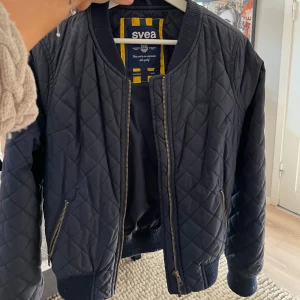 Svart quiltad bomberjacka från Svea - Snygg svart quiltad bomberjacka från Svea med dragkedja framtill och ribbade muddar. Perfekt för en stilren look.