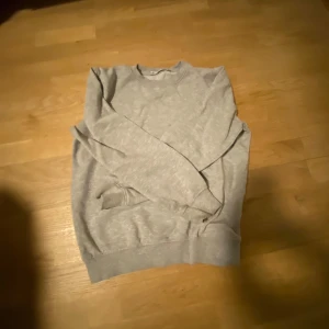 Grå sweatshirt - Säljer en klassisk grå sweatshirt med rund hals och långa ärmar. Perfekt för en avslappnad stil. Tröjan har ribbade muddar vid ärmslut och nederkant för extra komfort.