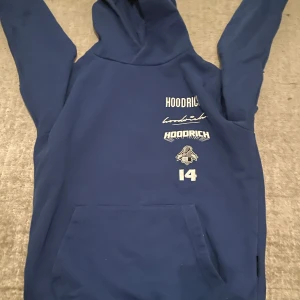 Blå hoodie från Hoodrich - Säljer en blå hoodie från Hoodrich med flera logotyper tryckta på framsidan och baksidan. Hoodien har en stor ficka framtill och en bekväm huva. Perfekt för en avslappnad stil.
