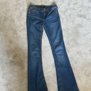 Blå bootcut jeans från True Religion - Snygga blå bootcut jeans från True Religion med klassisk femficksdesign och kontrastsömmar, rosa söm. Jeansen har en midwaist passform och är perfekta för en avslappnad stil. Märkeslogga på bakfickan ger en extra touch.