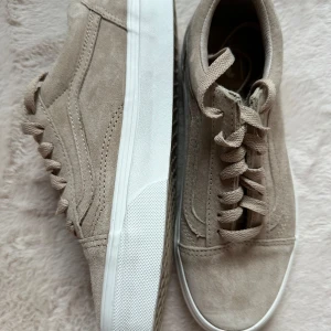 Beige sneakers från Vans - Säljer ett par snygga beige sneakers från Vans i beige mocka. Skorna har en klassisk design med snörning och en vit sula. Perfekta att matcha till klänning i sommar eller att ha på studenten! Storlek 36,5. Hämtas i Umeå, eller så står köparen för frakten!