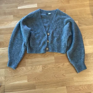 Grå stickad kofta med knappar - Säljer en mysig grå stickad kofta med stora knappar framtill. Koftan har en v-ringad design och långa ärmar, perfekt för kyligare dagar. Den är croppad och ger en trendig look. Perfekt för lager-på-lager-stil!