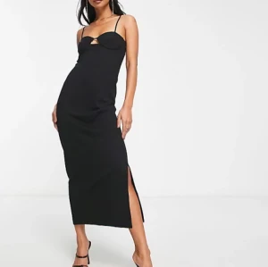 Svart tajt långklänning  - Elegant svart långklänning med smala axelband och en stilren slits på sidan. Klänningen har en unik detalj vid bysten som ger en modern touch. Perfekt för en kväll ute.