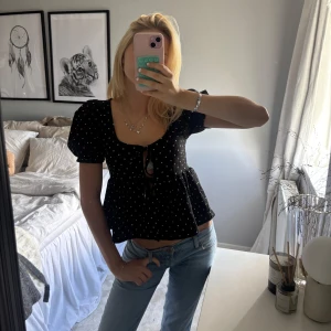 Svart prickig blus med puffärm - Supersöt svart blus med vita prickar och puffiga korta ärmar. Toppen har en söt knytning framtill och lätt utställd passform nedtill. Djup, fyrkantig ringning och luftig känsla – perfekt till jeans eller kjol.