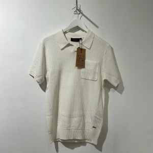 Offwhite pikétröja från Indicode - Snygg offwhite pikétröja från Indicode i ribbad bomull. Tröjan har klassisk krage, korta ärmar och en bröstficka som ger en clean look. Perfekt för dig som gillar stilrena och enkla plagg med lite struktur. 100 bomull 