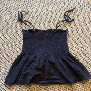 Mörkblå linnetopp  - Supersöt mörkblå linnetopp med smockad överdel och volangig nederdel. Axelbanden är tunna och knyts på axlarna. Perfekt för varma dagar och enkel att styla med jeans eller kjol.