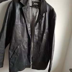 Svart skinnjacka från Patino, M - Klassisk svart skinnjacka från Patino Leather Company i storlek M. Jackan har en rak passform, krage och knäppning framtill. Två snedställda fickor och stilren design gör den enkel att matcha med olika outfits. Upplever den som stor i storlek, är nog en herrmodell. 