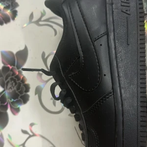 Svarta Nike Air Force 1 sneakers - Klassiska helsvarta Nike Air Force 1 sneakers i skinn med svart sula och svarta skosnören. Snygg och tidlös modell med perforerad tå och silverfärgade AF1-detaljer på snörningen. Perfekta för dig som gillar streetstyle och vill ha en ikonisk look.