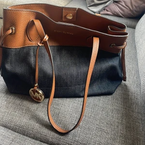 Michael Kors brun och svart axelväska - Snygg axelväska från Michael Kors i brunt skinn och svart jeansmaterial. Väskan har långa handtag, guldfärgad MK-detalj och praktiska innerfack med dragkedja. Perfekt för dig som vill ha en rymlig och stilren väska med exklusiv känsla.