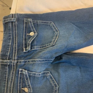 Blå jeans low waisted - Säljer ett par klassiska blå jeans med tydliga vita kontrastsömmar och coola fickdetaljer med lock och knapp. Jeansen är i fint skick. St 152 passar även uppåt 160!