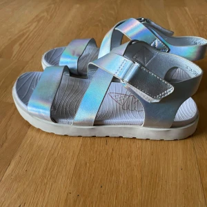 Silvriga sandaler med breda remmar - Snygga sandaler i skimrande silver med breda remmar och vit platt sula. Remmarna har en cool holografisk effekt och stängs med kardborre. Perfekta för sommardagar när du vill sticka ut lite extra.